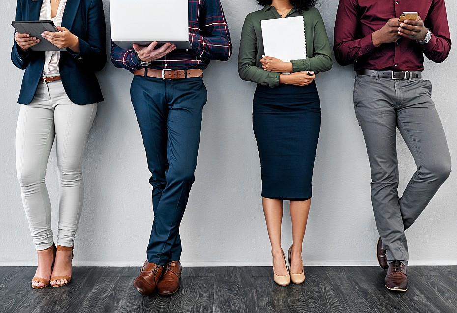 Vier Personen in Business-Kleidung stehen vor einer Wand und halten Laptop, Tablet, Smartphone und Block in der Hand