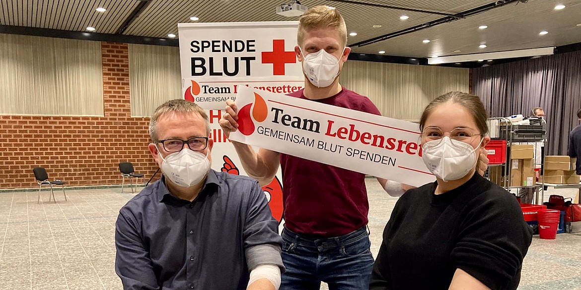 Drei Personen nach der Blutabnahme. Die mittlere Person hält einen "Team Lebensretter" Banner